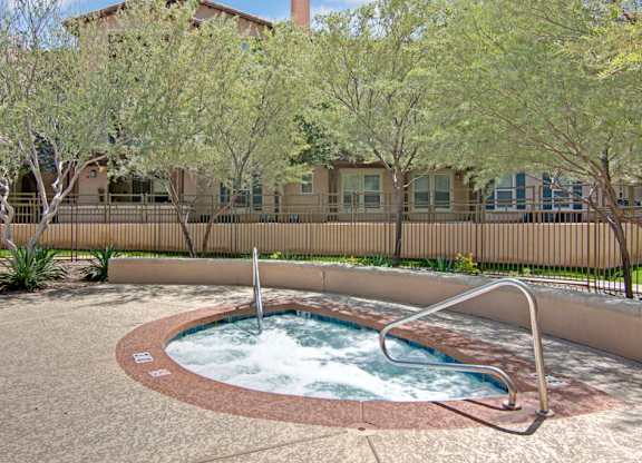 A relaxing spa area here at Bela Rosa featuring a bubbling in-ground hot tub surrounded by a spacious concrete deck and secure fencing. Lush desert landscaping and mature trees provide shade and privacy, while nearby apartment homes create a comfortable, resort-inspired setting perfect for unwinding after a long day.