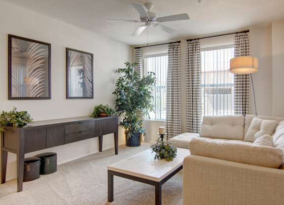 A bright and inviting living room here at Bela Rosa featuring plush neutral carpeting, a comfortable beige sofa, and a sleek coffee table centered beneath a ceiling fan. Large windows with patterned curtains bring in natural light, while framed wall art, a modern console table, and lush greenery add warmth and style to this cozy gathering space.