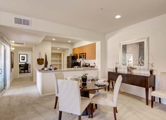 A spacious open dining and kitchen area here at Bela Rosa featuring plush carpeting, a round dining table with upholstered chairs, and a raised breakfast bar overlooking the kitchen. Light wood cabinetry, stainless steel appliances, and recessed lighting complement the airy layout, while a sliding glass door brings in natural light and extends the living space outdoors.