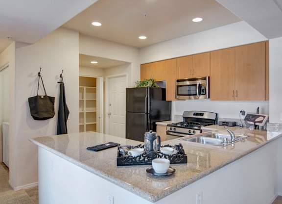 A bright open kitchen here at Bela Rosa featuring light wood cabinetry, granite-style countertops, and a spacious breakfast bar with an undermount sink. Stainless steel appliances and recessed lighting enhance the modern layout, while an adjacent hallway with built-in shelving adds convenient storage and seamless flow into the living areas.