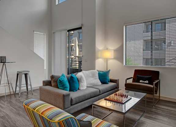 A bright two-story living room here at Escape featuring soaring ceilings, expansive windows with blinds, and wood-style flooring, styled with a gray sofa accented by teal and white pillows, a glass coffee table with a chess set, a cozy armchair, floor lamp, and sliding glass door leading to a private balcony that fills the space with natural light.