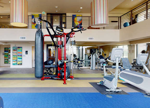 A spacious contemporary fitness center here at Escape featuring an open two-story layout with large windows, colorful pendant lighting, and a wide range of strength and cardio equipment including weight machines, stationary bikes, treadmills, and a dedicated boxing area, all set on cushioned flooring for comfortable, energizing workouts.