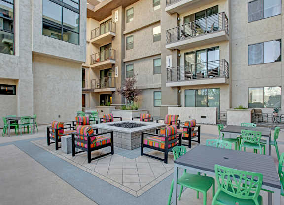 A modern outdoor courtyard here at Escape featuring a central fire pit surrounded by colorful cushioned lounge chairs, contemporary dining tables with green seating, tiled patio flooring, and sleek apartment balconies overhead, creating a vibrant and inviting space for gathering, relaxing, and enjoying Arizona’s sunny weather.