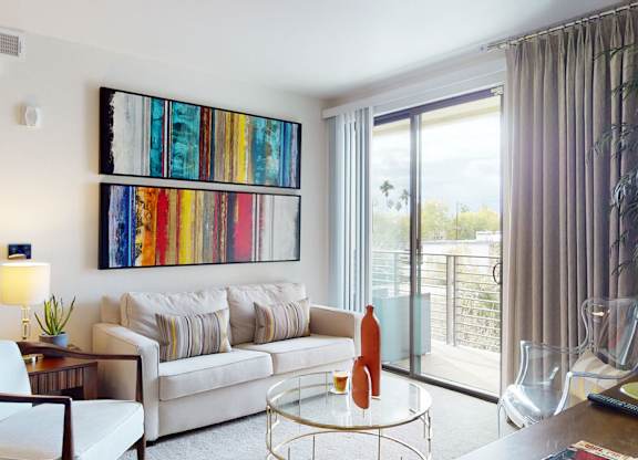 A bright contemporary living room here at Escape featuring a plush neutral sofa with patterned accent pillows, a round glass coffee table with decorative vases, and colorful abstract wall art, complemented by a cozy armchair and side table with lamp, while a sliding glass door with full-length curtains opens to a private balcony overlooking scenic views and palm trees.