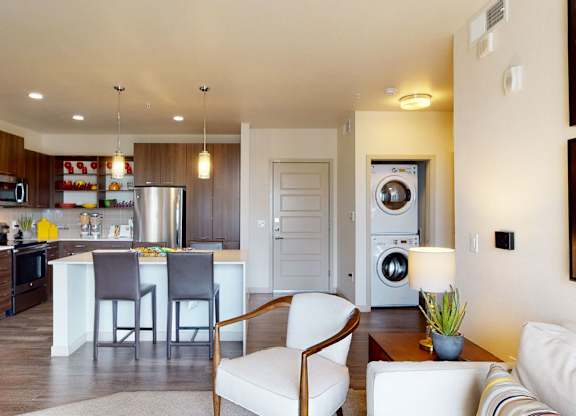 A bright open-concept living and kitchen space here at Escape showcasing warm wood-style cabinetry, stainless-steel appliances, and a spacious island with bar seating under modern pendant lighting, complemented by wood-style flooring, a cozy living area with accent chair and sofa, and a convenient in-home stacked washer and dryer tucked neatly beside the entry door.