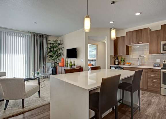 A contemporary open kitchen and living area here at Escape featuring rich wood-style cabinetry, stainless-steel appliances, and a sleek quartz island with bar seating under modern pendant lighting, complemented by wood-style flooring, soft carpeting in the living space, vertical blinds leading to a private balcony, and stylish furnishings that create a warm, inviting atmosphere.