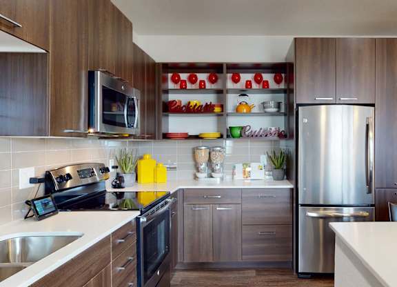 A sleek modern kitchen here at Escape featuring rich wood-style cabinetry with brushed metal hardware, stainless-steel appliances, a smooth-top range, built-in microwave, and spacious refrigerator, complemented by quartz countertops, a tile backsplash, open display shelving with decorative accents, and an expansive island that creates a stylish and functional cooking space.