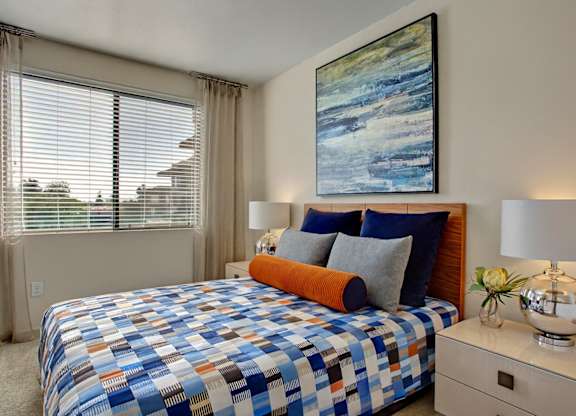 A stylish bedroom here at Escape featuring a warm wood headboard, layered blue and gray bedding with an accent bolster pillow, and matching nightstands topped with modern chrome lamps, while a large window with blinds and soft drapery fills the space with natural light and a bold abstract artwork adds a contemporary focal point above the bed.