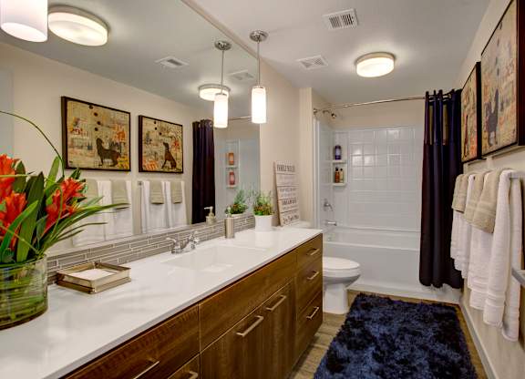 A contemporary bathroom here at Escape featuring a long wood-style vanity with sleek hardware, a wide mirror with modern lighting above, and a spacious white countertop with integrated sink, while a full tub and shower combination with tile surround, dark curtain accents, plush bath rug, and neatly hung towels create a polished and inviting retreat.