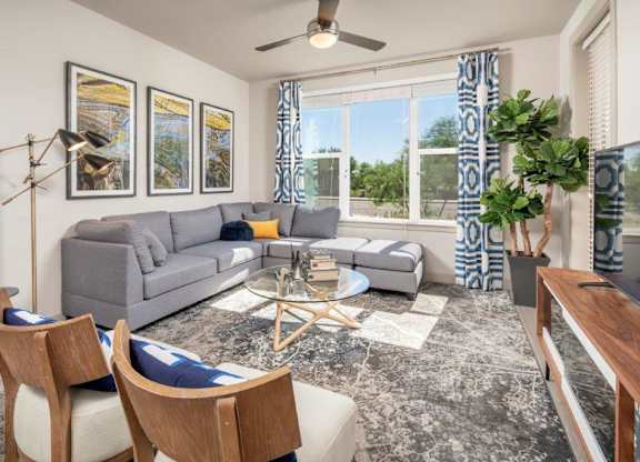 A bright and contemporary living room here at The Flats at San Tan featuring a spacious gray sectional sofa with accent pillows, three framed art pieces, and a sleek floor lamp, complemented by a round glass coffee table on a patterned area rug, large windows with blue geometric curtains, a ceiling fan, and a stylish media console creating an inviting and airy atmosphere.