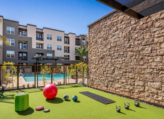 A vibrant outdoor fitness area here at The Flats at San Tan featuring green turf flooring with kettlebells, weighted balls, a yoga mat, and stacked step platforms, set beside a stone accent wall and overlooking the resort-style pool and modern apartment buildings with palm trees and private balconies.