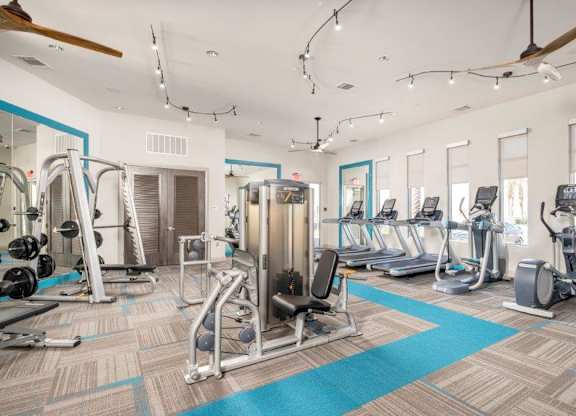 A spacious fitness center here at The Flats at San Tan featuring strength-training machines, free weights, and multiple cardio options including treadmills and elliptical machines, all arranged on patterned flooring with teal accents, large windows for natural light, ceiling fans, and modern lighting for an energizing workout environment.