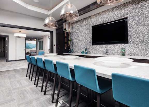 A modern entertainment lounge here at The Flats at San Tan featuring a long white quartz bar-height table with teal upholstered stools, pendant lighting overhead, a textured accent wall with a mounted flat-screen television, and sleek tile flooring, creating a stylish space for residents to gather, work, or socialize.