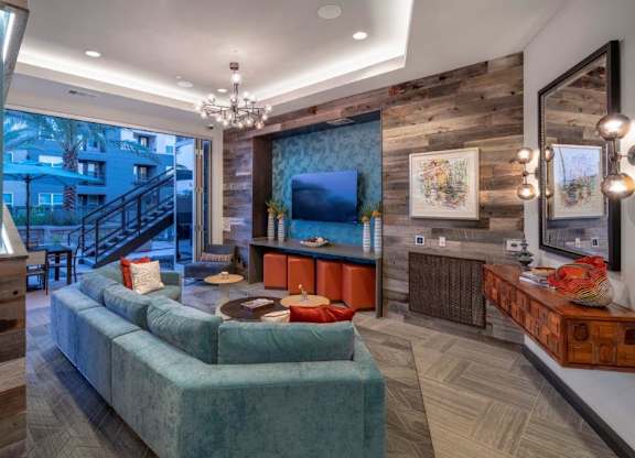 A stylish indoor lounge here at The Flats at San Tan featuring a plush teal sectional sofa with accent pillows, round wood coffee tables, and a built-in media wall with mounted television and wood plank detailing, complemented by modern globe lighting, textured accent walls, and sliding glass doors that open to an inviting outdoor courtyard space.