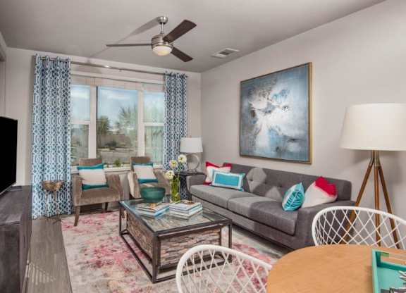 A stylish and inviting living room here at The Flats at San Tan featuring a gray sofa with vibrant accent pillows, two upholstered armchairs, and a glass-top coffee table with a stone base set atop a soft patterned rug, complemented by large windows with blue geometric curtains, a modern ceiling fan, contemporary artwork, and a sleek media console for a bright, welcoming feel.