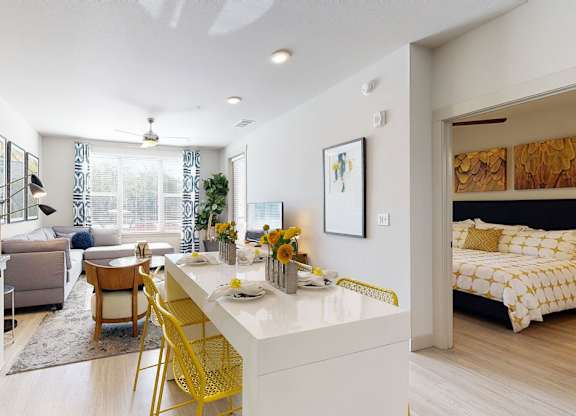 A bright and contemporary open-concept living and dining area here at The Flats at San Tan featuring a sleek white kitchen island with modern yellow barstools, granite countertops, and stainless steel appliances, flowing into a cozy living room with a plush sectional, patterned rug, and large windows with blue curtains, while a spacious bedroom with coordinated bedding is visible just beyond.
