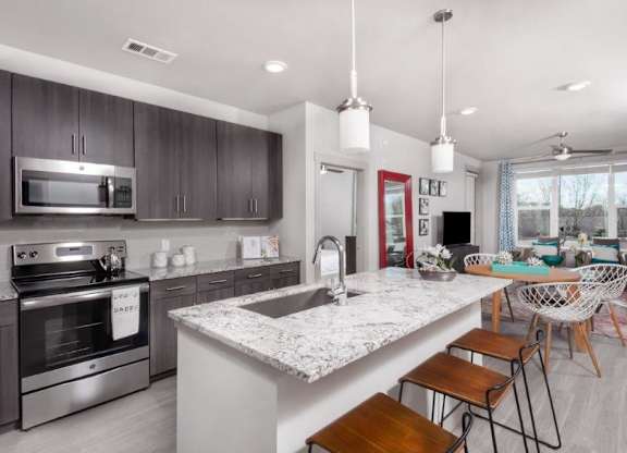 A sleek and inviting open kitchen and dining space here at The Flats at San Tan featuring dark wood cabinetry, stainless steel appliances, and a spacious granite island with bar seating, illuminated by modern pendant lighting, flowing seamlessly into a bright living area with large windows, contemporary furnishings, and light wood-style flooring for a clean, airy feel.