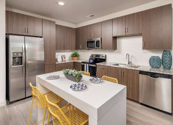 A bright and contemporary kitchen here at The Flats at San Tan featuring rich wood cabinetry, sleek stainless steel appliances, and light granite countertops paired with a white tile backsplash, highlighted by a spacious island with vibrant yellow seating, modern fixtures, and wood-style flooring that create a clean, welcoming space ideal for cooking and gathering.