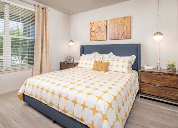 A serene and stylish bedroom here at The Flats at San Tan featuring a plush upholstered bed dressed in white bedding with golden patterned accents, complemented by matching nightstands, warm wood finishes, and modern pendant lighting, while a large window with soft curtains brings in natural light to create a bright and inviting retreat.