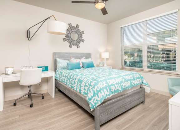 A bright and contemporary bedroom here at The Flats at San Tan featuring a gray wood bed frame dressed in vibrant teal and white patterned bedding, accented by coordinating pillows and a decorative wall mirror above the headboard, while a built-in desk with a modern task chair and a large window with blinds fill the space with natural light and functionality.