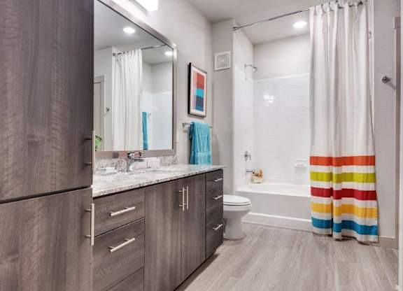 A bright and modern bathroom here at The Flats at San Tan showcasing wood-style cabinetry with sleek hardware, a spacious granite countertop with an undermount sink, and a large framed mirror that enhances the open feel, complemented by wood-look flooring, a white tub and shower combination, and a colorful striped shower curtain that adds a vibrant touch to the space.