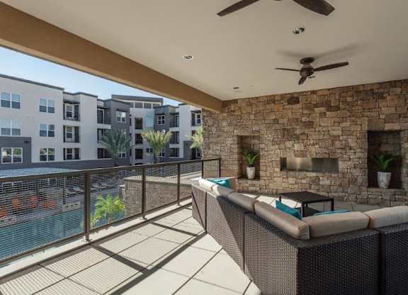 A spacious covered patio lounge here at The Flats at San Tan featuring woven sectional seating with plush cushions, a sleek outdoor coffee table, and a striking stone accent wall with a built-in fireplace, complemented by ceiling fans, recessed lighting, and a metal railing overlooking the sparkling pool and surrounding apartment homes for an inviting community gathering space.