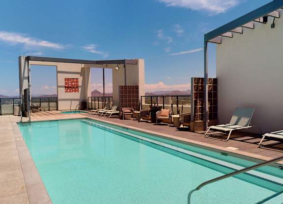 A vibrant rooftop pool deck here at JOY on 4th showcasing a long, resort-style swimming pool surrounded by sleek lounge chairs and shaded pergola structures. Glass railings frame expansive mountain and city views, while comfortable seating areas create inviting spaces to unwind, socialize, and enjoy Arizona sunshine in a modern urban setting.