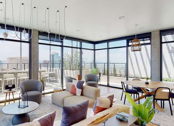 A panoramic rooftop lounge here at JOY on 4th showcasing floor-to-ceiling windows with sweeping skyline and mountain views, contemporary seating in soft neutral tones, and warm wood flooring. Stylish armchairs and a plush sofa surround modern tables, while a dining area and outdoor terrace seating create an open, light-filled space ideal for relaxing or gathering.