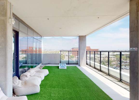 A modern rooftop lounge here at JOY on 4th featuring lush green turf flooring, relaxed bean bag seating along a glass wall, and sleek concrete columns framing open skyline views. Glass railings overlook the city, while shaded overhead coverage creates a breezy, elevated retreat ideal for casual gatherings and outdoor relaxation.
