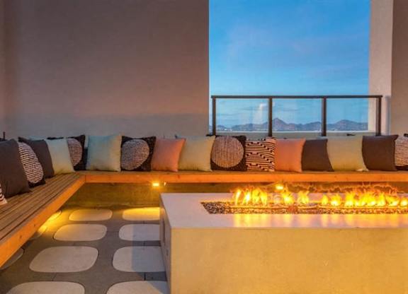 A cozy rooftop lounge here at JOY on 4th featuring built-in wood bench seating with plush accent pillows surrounding a sleek fire pit table. Warm ambient lighting and patterned pavers create an inviting atmosphere, while glass railings frame stunning sunset views of the city skyline and distant mountains for a relaxing evening retreat.