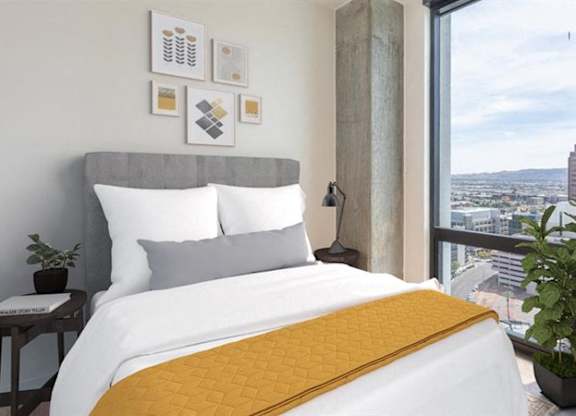 A bright modern bedroom here at JOY on 4th featuring a plush upholstered headboard, crisp white bedding with a textured mustard accent throw, and minimalist wall art above. A sleek nightstand with a contemporary lamp sits beside the bed, while floor-to-ceiling windows showcase sweeping city and mountain views and fill the space with natural light.