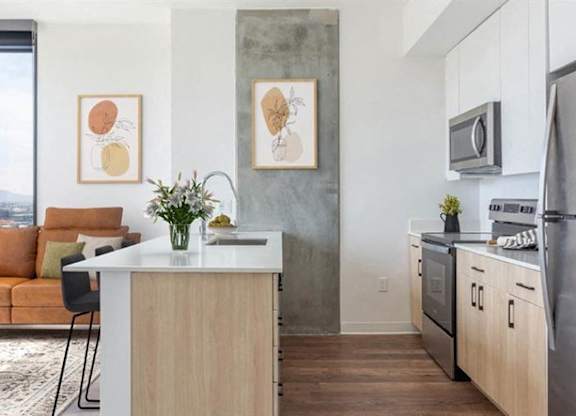 A bright contemporary living and kitchen space here at JOY on 4th featuring a sleek island with light wood cabinetry, white quartz countertops, and stainless-steel appliances. A warm-toned sofa and modern artwork add inviting style, while floor-to-ceiling windows frame expansive city and mountain views and fill the open layout with natural light.