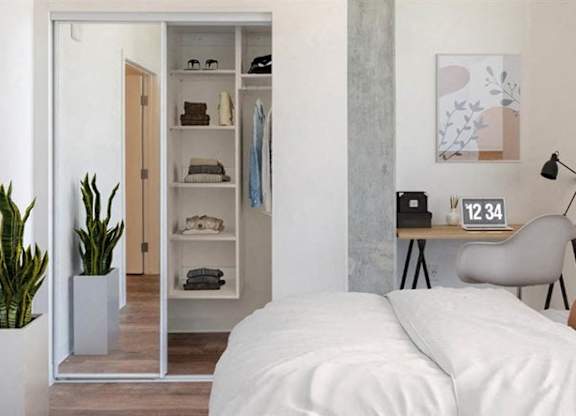 A bright modern bedroom here at JOY on 4th featuring soft neutral bedding, a warm accent pillow, and a sleek wall-mounted reading lamp above a compact desk workspace. Sliding mirrored closet doors reveal built-in shelving, while a tall potted plant and contemporary artwork add inviting style to this light-filled retreat.