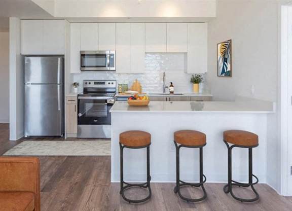 A bright contemporary kitchen here at JOY on 4th showcasing sleek white cabinetry, stainless steel appliances, and a spacious peninsula with three upholstered barstools. Light wood flooring and a glossy tile backsplash add warmth and texture, while an open layout connects seamlessly to the living area for modern, functional living.