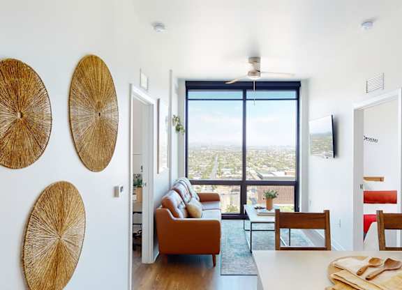 A sleek and inviting living area here at JOY on 4th featuring expansive floor-to-ceiling windows with sweeping city views, warm wood-style flooring, and a contemporary orange sofa that anchors the space, complemented by modern wall art, a streamlined ceiling fan, and an open layout that connects seamlessly to the kitchen and dining area for comfortable urban living.