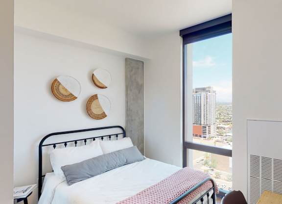 A bright and modern bedroom here at JOY on 4th featuring a sleek black metal bed frame with crisp white bedding and a soft accent throw, complemented by woven wall décor and a concrete accent column. Expansive floor-to-ceiling windows showcase sweeping city views while natural light enhances the clean lines, warm wood flooring, and contemporary finishes.