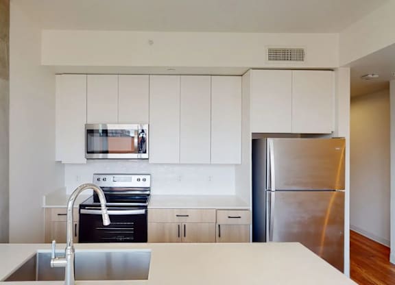 A sleek and contemporary kitchen here at JOY on 4th featuring crisp white upper cabinetry paired with warm wood lower cabinets and streamlined black hardware. Stainless steel appliances, a smooth tile backsplash, and expansive quartz-style countertops create a clean, modern aesthetic, while the spacious island with an undermount sink offers ample room for cooking, dining, and entertaining.