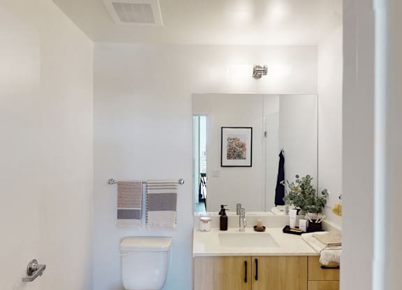 A bright and minimalist bathroom here at JOY on 4th featuring a sleek vanity with warm wood cabinetry and modern black hardware paired with a clean white countertop and integrated sink. A large frameless mirror enhances the open feel, while polished fixtures, neatly arranged towels, and simple greenery accents create a fresh, contemporary space designed for comfort and functionality.