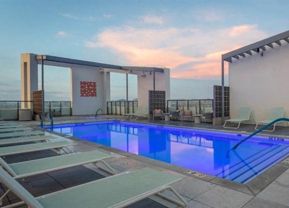 A stunning rooftop pool deck here at JOY on 4th featuring a sleek rectangular swimming pool illuminated with vibrant blue lighting and surrounded by modern lounge chairs. Framed by clean architectural lines and glass railings, this elevated retreat offers sweeping city views, open sky sunsets, and stylish seating areas designed for relaxing, socializing, and soaking in the urban atmosphere.