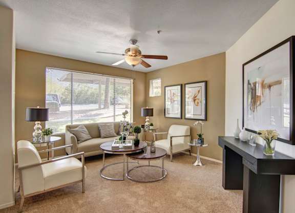 A cozy inviting living room here at Mirador featuring warm beige walls, plush carpet flooring, and a large window with blinds that fills the space with natural light. A neutral sofa and two modern accent chairs surround nested coffee tables, while sleek side tables, table lamps, and framed artwork create a comfortable, welcoming setting for everyday relaxation.
