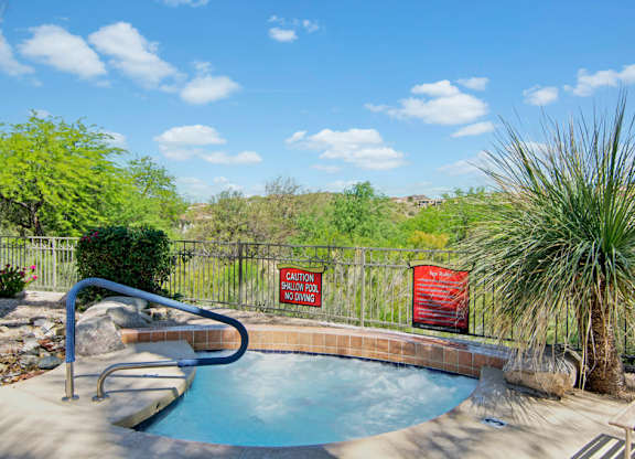 A relaxing outdoor spa here at Mirador featuring a circular hot tub with tiled surround and curved stainless handrail, set against desert landscaping and scenic hillside views. Wrought iron fencing, natural stone accents, and nearby lounge seating create a peaceful retreat ideal for unwinding under sunny Arizona skies.