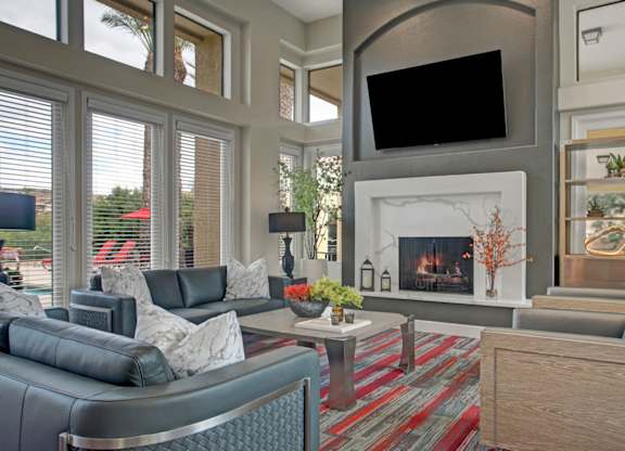 A welcoming clubhouse living room here at Mirador featuring soaring ceilings and expansive windows that fill the space with natural light. Contemporary leather seating surrounds a sleek coffee table and modern fireplace, while a mounted television and built-in shelving create a stylish, comfortable setting for relaxing or gathering with neighbors.
