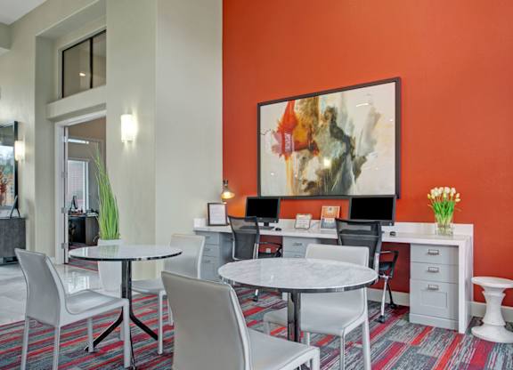 A modern business center here at Mirador featuring a bold red accent wall with large abstract artwork, built-in workstations with computers and ergonomic chairs, and sleek marble-top tables for collaborative seating. Neutral tones and contemporary lighting create a bright, professional space ideal for remote work or study.