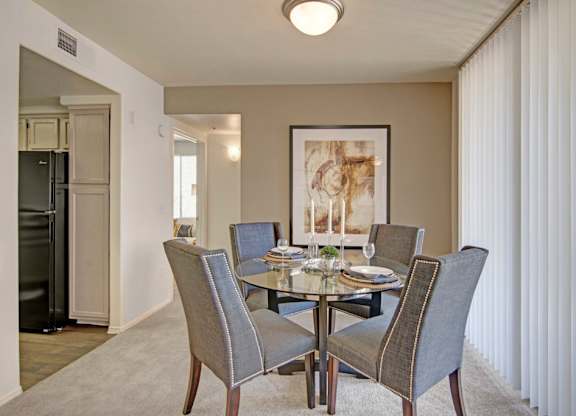 A warm welcoming dining area here at Mirador featuring plush carpet flooring and a round glass-top table surrounded by four upholstered chairs with nailhead trim. A large framed art piece anchors the space, while vertical blinds filter natural light and an open entryway leads toward the kitchen, creating an inviting setting for meals and gatherings.