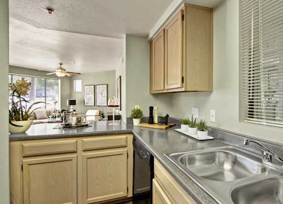 A functional kitchen here at Mirador featuring light wood cabinetry, gray countertops, and a double-basin stainless sink beneath a window with white blinds. The open layout flows into the adjacent living area, while decorative accents, small plants, and ample counter space create a bright and practical setting for everyday cooking and entertaining.