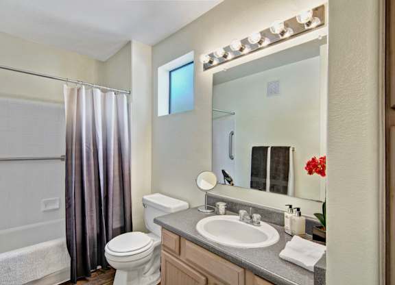 A well-lit bathroom here at Mirador showcasing a spacious vanity with light wood cabinetry, a wide mirror with bright overhead lighting, and a smooth countertop with an integrated sink. A full tub and shower combination with tile surround and curtain sits beside a frosted window, while wood-style flooring adds warmth to the space.