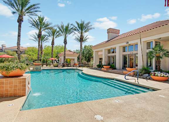 A resort-style swimming pool here at Mirador surrounded by tall palm trees, terracotta tile accents, and spacious sun decks. The sparkling blue water curves alongside wide entry steps and stainless handrails, while vibrant planters, shaded seating areas, and a stucco clubhouse with red tile roofing create a warm, inviting atmosphere perfect for relaxing outdoors.