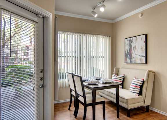 A cozy dining nook here at Mozaic at Steele Park featuring wood-style flooring, a modern table with seating for four, and a cushioned bench with accent pillows. Large windows with vertical blinds and a nearby glass door provide abundant natural light, creating a bright and inviting space for everyday meals or entertaining.