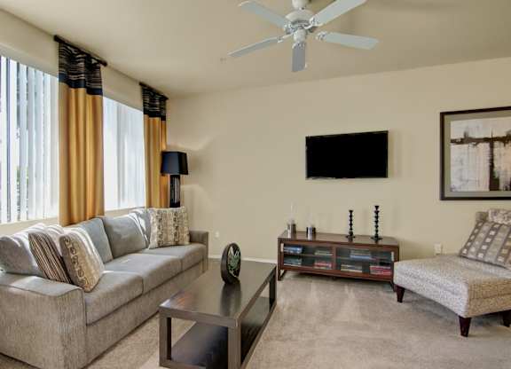 A cozy living room here at Natura Villas featuring a plush neutral sofa accented with patterned throw pillows, a coordinating lounge chair, and a sleek dark wood coffee table. Large windows with vertical blinds and drapery panels allow natural light to brighten the space, while a mounted television and low media console create a comfortable setting for relaxing or entertaining.