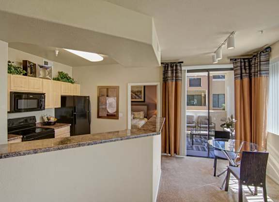 A bright open-concept kitchen and dining space here at Natura Villas featuring light wood cabinetry, expansive countertop space, and modern appliances. The breakfast bar overlooks a cozy dining nook with a glass-top table, while sliding glass doors lead to a private patio and allow natural light to fill the room, creating a comfortable and connected living environment.